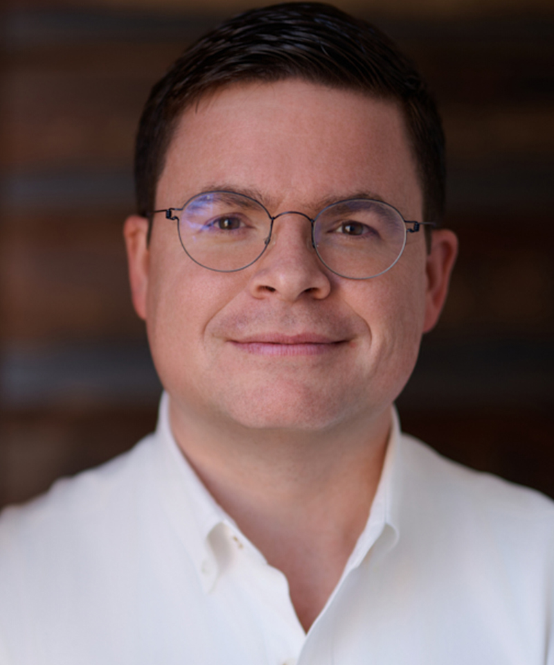 Headshot photo of Jimmy Goodrich, a white man wearing glasses in a white dress shirt in front of a brown background