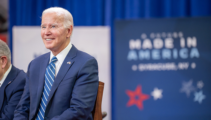 President Joe Biden smiling in a blue suit, in front of a blue MADE IN AMERICA poster with red, white, and blue stars.