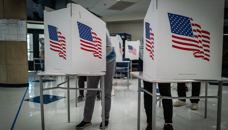 Two white voting booths with American flags on them. Two people are hidden behind the voting booth.