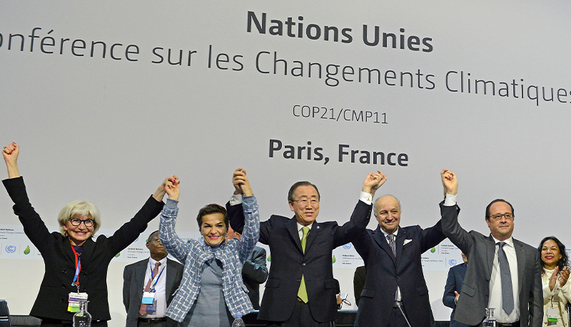 A group of world leaders at COP 21 after the signing of the Paris Agreement. They are holding their joined hands in the air in triumph against a white background.