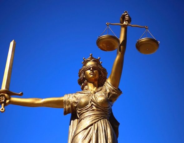 A bronze sculpture depicting Lady Justice, a blindfolded woman holding scales in one hand and a sword in the other, against a blue background.