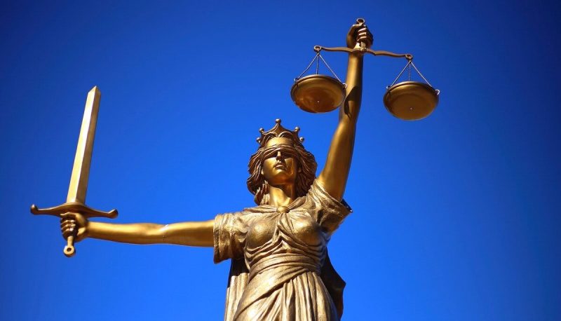 A bronze sculpture depicting Lady Justice, a blindfolded woman holding scales in one hand and a sword in the other, against a blue background.