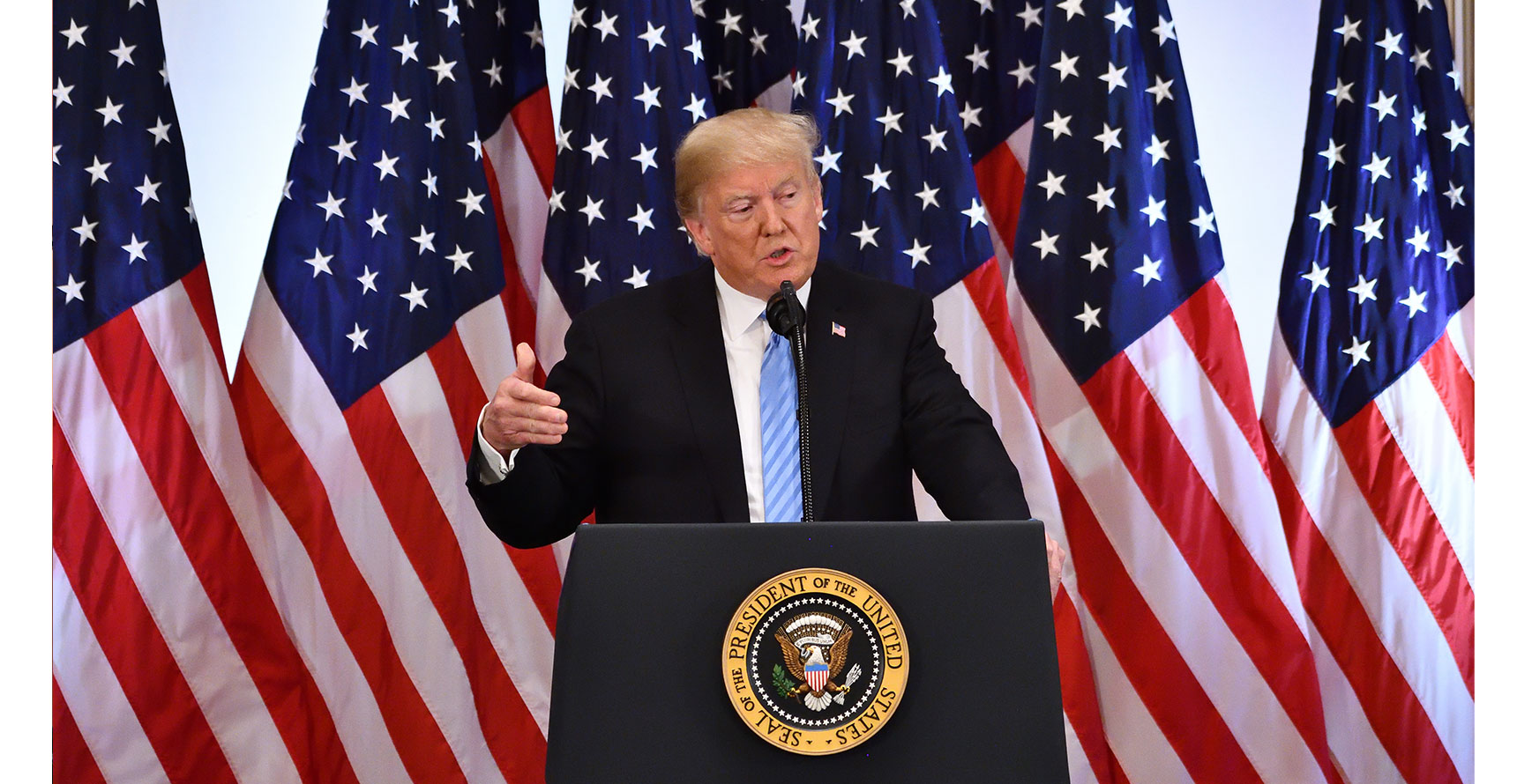 President Donald Trump speaks at a podium in front of a backdrop of several American flags.