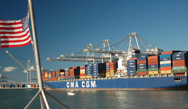 An image of a cargo ship transporting multiple colorful shipping containers. An American flag waves in the foreground.