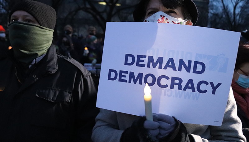 A photo of a protestor holding a sign that reads "Demand Democracy"
