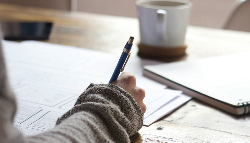 Close-up photo of a person writing on a blank piece of paper with a mug nearby