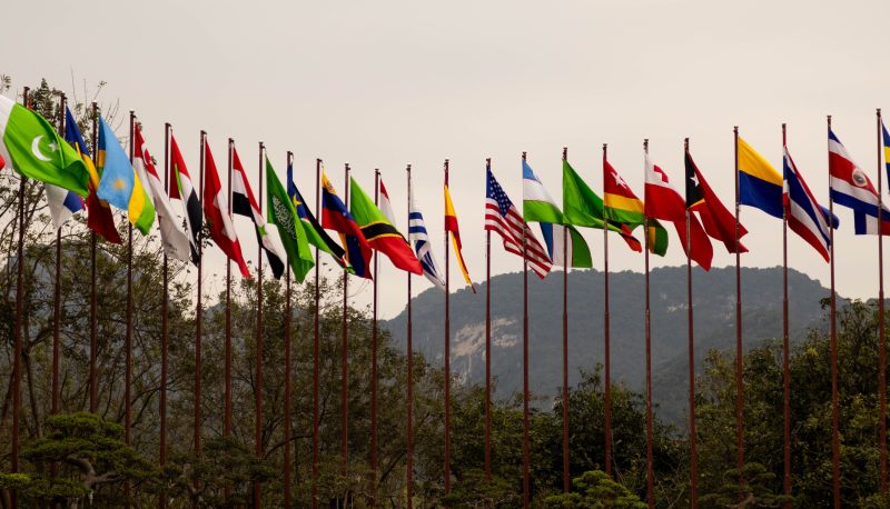 A display of various national flags on flagpoles set against a mountain backdrop, symbolizing global unity.